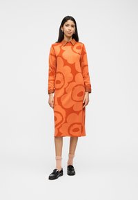 Γυναίκα που στέκεται, φοράει ένα πορτοκαλί μεσοφόρι με μακριά μανίκια και μεγάλα λουλουδένια σχέδια, γιακά, ροζ κάλτσες και μαύρα μοκασίνια.