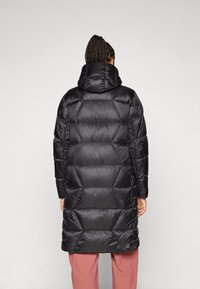 Osoba u crnom pufferskom kaputu s kapuljačom i pletenicom, viđena s leđa, stoji pred svijetlom pozadinom.