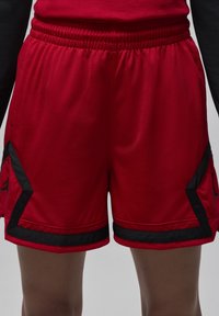 Punaiset urheilushortsit hengittävästä materiaalista. Niissä on mustia yksityiskohtia ja sivupaneelit, joissa on logo. Mukavuuden takaamiseksi niissä on joustava vyötärönauha.