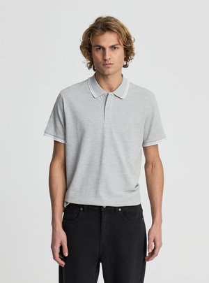 Giovane uomo con capelli ricci biondi che indossa una polo grigio chiaro a maniche corte e pantaloni neri, in piedi davanti a uno sfondo chiaro e uniforme.