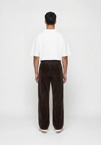 Άνδρας στέκεται με την πλάτη γυρισμένη, φορώντας λευκό πουκάμισο με κοντά μανίκια, σκούρο καφέ παντελόνι κορντόρι και λευκά αθλητικά παπούτσια, σε απλό φόντο.