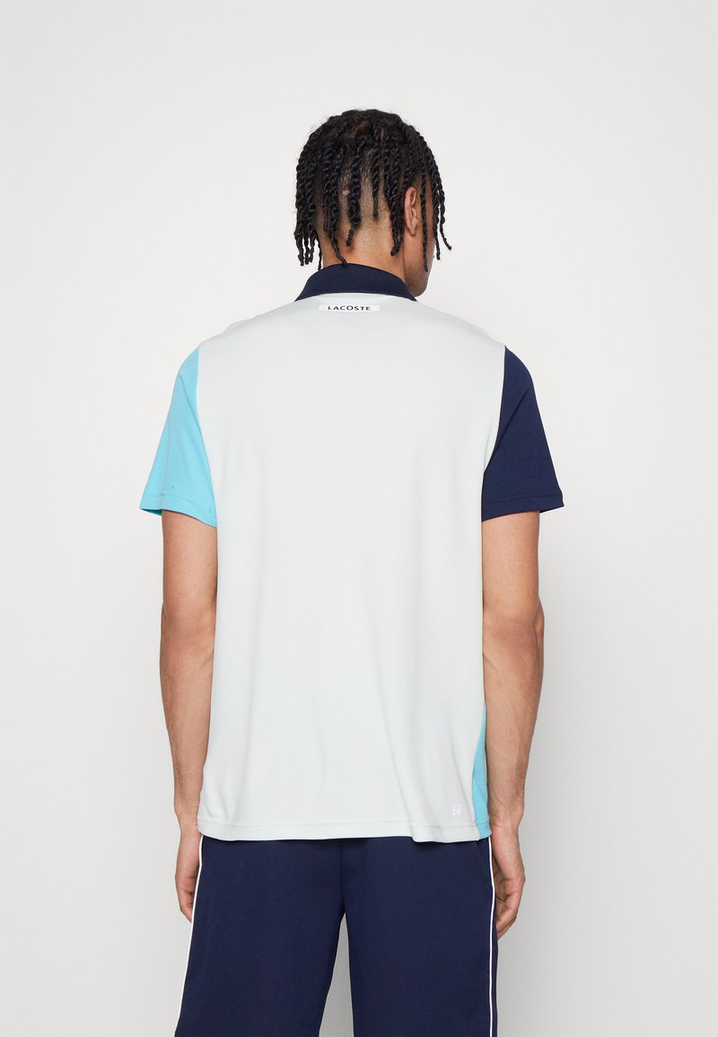 Polo-shirt avec un design coloré en bloc comprenant des sections gris clair, teal et marine, à manches courtes et avec un col. Logo Lacoste à l'arrière.