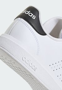 Zapatilla blanca con acento de cuero negro en el talón, suela texturizada, diseño perforado en los lados y prominente logo "adidas" en el talón.