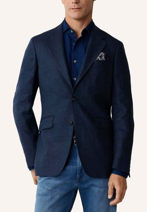 Man draagt een donkerblauw geruit colbert met twee knopen, een marineblauw overhemd, een pochet en een blauwe spijkerbroek, staand tegen een effen achtergrond.