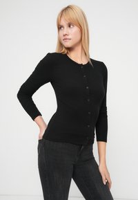 Cardigan negru cu dungi, cu decolteu rotund și mâneci lungi, prevăzut cu nasturi negri pe partea din față. Materialul pare moale și care se mulează pe corp.