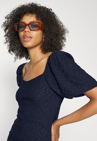Vestido azul marinho com mangas bufantes e detalhes em ilhós. Modelo usa óculos com lentes laranja. Textura suave e decote quadrado.