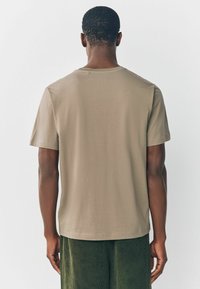 Rövid ujjú, bézs színű póló, puha anyagból. Klasszikus kerek nyakú dizájn, egyenes hem. Texturált zöld nadrággal viselve.