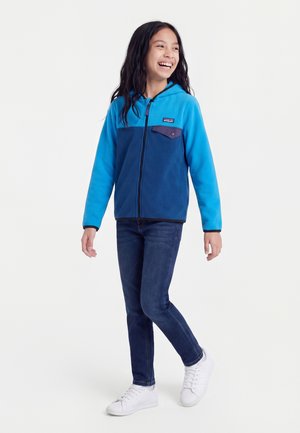 Fille aux longs cheveux foncés souriante, portant un sweat à capuche zippé bleu à blocs de couleur, un jean foncé et des baskets blanches, debout devant un fond uni.