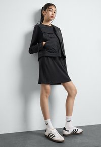 Vestido preto canelado combinado com um colete preto com fecho éclair à frente e com bolsos. Ténis brancos com riscas pretas e logótipo, complementados por meias brancas.