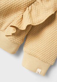 Tessuto giallo con texture, orlo arricciato e polsini lavorati a maglia. Include un piccolo cartellino con la lettera 'A' stampata, che enfatizza i dettagli del design.
