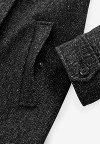 Detaliu al unui palton din lână gri, cu model herringbone, care include un buzunar lateral, manșete cu nasturi și o țesătură texturată cu un model subtil.