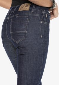 Gros plan sur un jean en denim bleu foncé avec des coutures marron clair, une poche arrière, une étiquette de marque brune, et la main d'une personne posée sur sa hanche.
