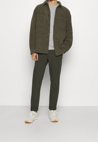 Olivově zelená texturovaná košile, šedé tričko s kulatým výstřihem, tmavě zelené slim-fit kalhoty a béžové běžecké boty s oranžovými akcenty.