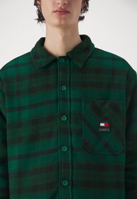 Camicia in flanella a quadri verdi e neri con bottoni sulla parte frontale, colletto, taschino sul petto e una patch con logo sulla tasca. Tessuto morbido.