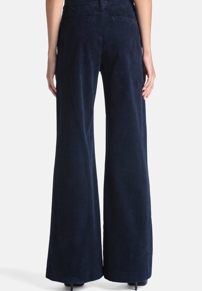 Femme portant un pantalon en velours côtelé bleu foncé taille haute avec jambes larges, vue de dos sur un fond blanc uni.