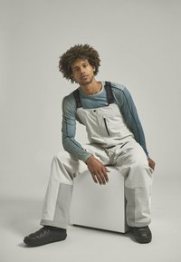 Weiße Latzhosen mit schwarzen Trägern, kombiniert mit einem türkisfarbenen Langarmshirt. Graue Akzente an den Unterschenkeln und schwarze Schuhe vervollständigen das Outfit.