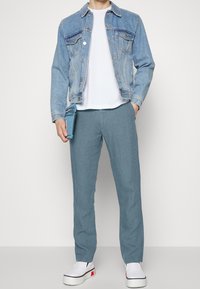Hellblaue Jeansjacke, weißes T-Shirt und hellblaue Leinenhose. Weiße Slipper mit roten Akzenten. Horizontale Struktur auf der Hose.