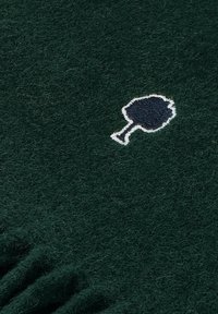 Groene wollen stof met een zachte textuur, voorzien van een geborduurd boompictogram in wit en marineblauw. Gerimpelde randen aan de onderkant.