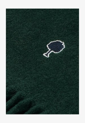 Grüner Wollstoff mit einer weichen Textur, der ein gesticktes Emblem eines Baumes in Weiß und Marineblau zeigt. Rüschenkanten entlang der Unterseite.