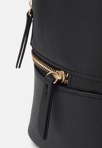 Sac noir texturé en matière synthétique, doté d'une fermeture éclair dorée bien visible avec un onglet décoratif. Fini lisse avec des accents minimaux.