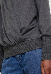 Ciemnoszary pulower z teksturowanym, prążkowanym ściągaczem w talii i przy mankietach, wykonany z miękkiego, gładkiego materiału o luźnym kroju. Połączony z niebieskimi dżinsami.