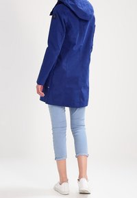 Blå vattentät jacka med huva, långa ärmar och rak skärning; har en logotyp på axeln, matchad med ljusblå jeans och vita skor.