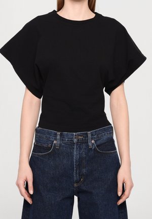 Kobieta ubrana w czarny krótki top z szerokimi rękawami i wysokim stanem ciemnoniebieskich dżinsów, stojąca na tle jednolitej ściany.