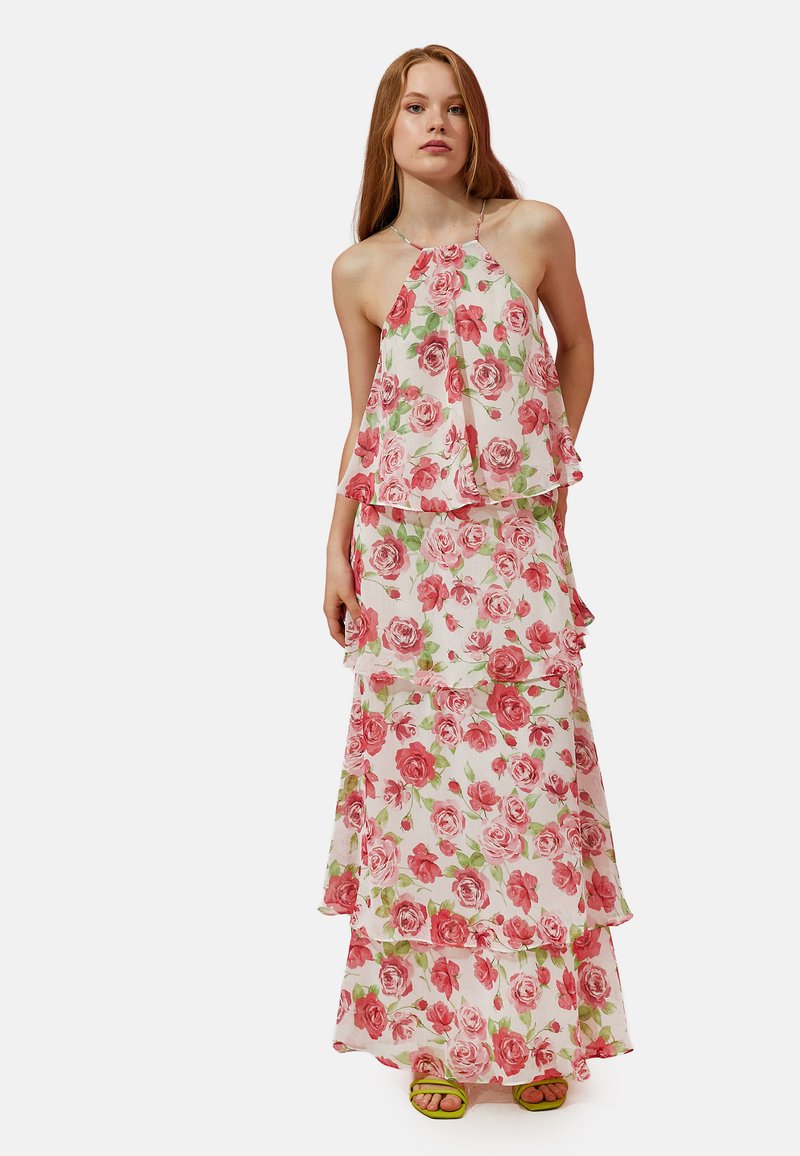 Maxikleid mit floralen Mustern und geschichteten Rüschen, das ein rosa-weißes Rosenmuster präsentiert. Hergestellt aus leichtem Stoff. Grüne Sandalen ergänzen den Look.