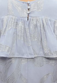 Ανοιχτό μπλε ύφασμα με σχέδια από ασημένια μεταλλική κλωστή, με διαφανές ραμμένο βολάν και τρία κουμπιά καλυμμένα με ύφασμα σε κλειστή πλάτη.