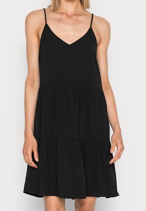 Femme portant une robe noire sans manches à volants, avec de fines bretelles et un décolleté en V, debout devant un fond clair uni.
