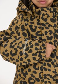 Barn iført en brun og sort leopardprintjakke med hætte, hånden stukket ind i frontlommen, og teksten "W-PRO 10000" på ærmet.