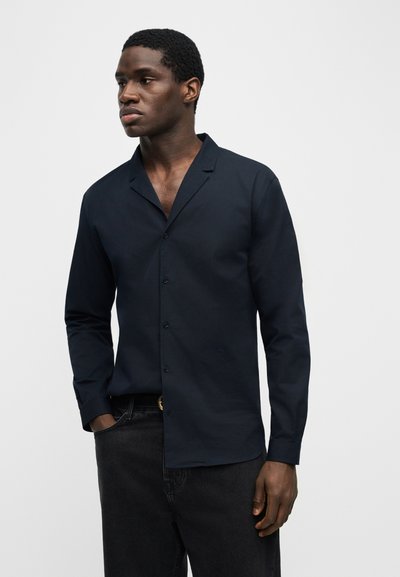 Mann trägt ein dunkelblaues Hemd mit Knöpfen und langen Ärmeln sowie schwarze Jeans, steht mit einer Hand in der Tasche vor einem schlichten Hintergrund.