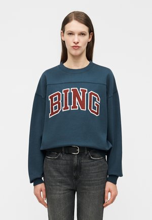 Γυναίκα που φοράει μια πετρόλ φούτερ με τη λέξη "BING" σε μεγάλα κόκκινα γράμματα, συνδυασμένη με μαύρη ζώνη και ξεθωριασμένα μαύρα τζιν, στέκεται μπροστά σε απλό φόντο.