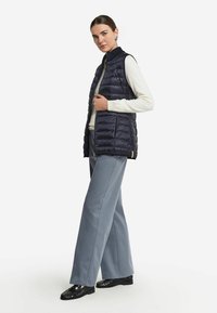 Marinblå väst i pufferstjärning med horisontell sömmar, hög krage och dragkedja. Matcher med ljusgrå vida byxor och svarta skor.