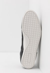 Black sneaker with a smooth upper, featuring a textured white rubber sole with circular patterns. Includes a Lacoste logo on the sole's side.