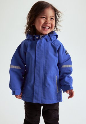 Smilende barn iført en blå vandtæt jakke med reflekterende striber på ærmerne og sorte bukser, stående foran en ensfarvet baggrund.