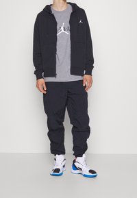 Person som bär svart zip-up hoodie, grå t-shirt med logotyp, svarta byxor och vita sneakers med blå och svarta accenter som står mot en vit bakgrund.