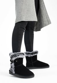 Botas negras hasta el tobillo con ribete de piel sintética, suelas planas y un detalle de hebilla plateada en contraste. Fabricadas en un material suave y texturizado.