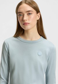 Helleblaue Langarmbluse mit Rundhalsausschnitt und strukturiertem Finish. Verfügt über ein kleines, tonales Logo-Patch im Brustbereich.
