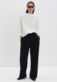 Donna che indossa occhiali da sole ovali neri, un maglione bianco largo, pantaloni neri a gamba larga e scarpe nere, in piedi con le mani in tasca su uno sfondo bianco.