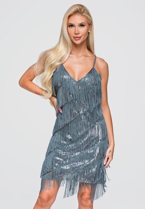 Femme blonde portant une mini-robe bleue sans manches avec franges et sequins, posant avec une main sur la hanche sur un fond neutre.