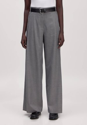 Person wearing high-waist black-and-white checkered wide-leg trousers with black belt and black shoes, standing against plain background.