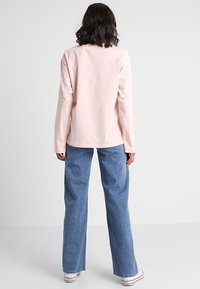 Person med skulderlangt hår iført en lys pink langærmet skjorte, blå vidde jeans og hvide sneakers, stående med ryggen til.