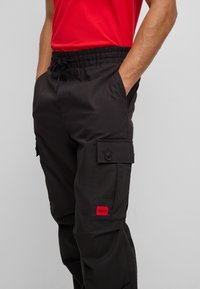 Black cargo pants featuring a drawstring waist, side pockets, and a red brand label. Fabric appears smooth with a subtle texture.
