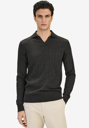 Mies pukeutunut tummanharmaaseen ribbineuletakkiin, jossa on kaulus ja pitkä hiha, sekä vaaleanbeigeihin housuihin, seisoo yksinkertaista valkoista taustaa vasten.