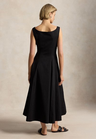 Kvinne med kort blondt hår iført en ermeløs, ankel-lang svart kjole og svarte sandaler, stående mot en beige bakgrunn.