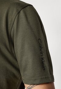 Alyvuogės žalios spalvos polo marškiniai su tekstūruotu audiniu, trumpomis rankovėmis ir juodu "Calvin Klein" logotipu, atspausdintu ant rankovės.