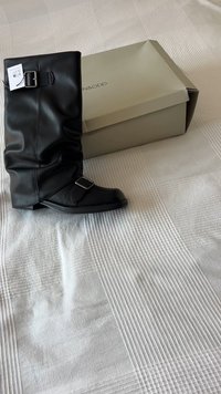 Botim alto de imitação de couro preto com design soltinho, tiras ajustáveis com fivela prateada, sola plana e bico arredondado, ao lado de uma caixa de sapatos.