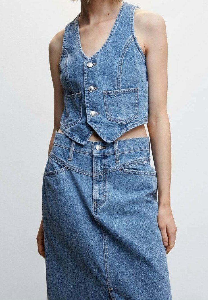 Femme portant un gilet en denim bleu sans manches avec des boutons et des poches avant, assorti à une jupe en denim bleu taille haute sur un fond uni.