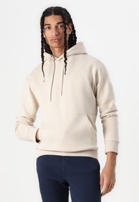 Beige hættetrøje lavet af blødt stof, med en frontlomme, snøre og ribbede manchetter. Båret med marineblå bukser.